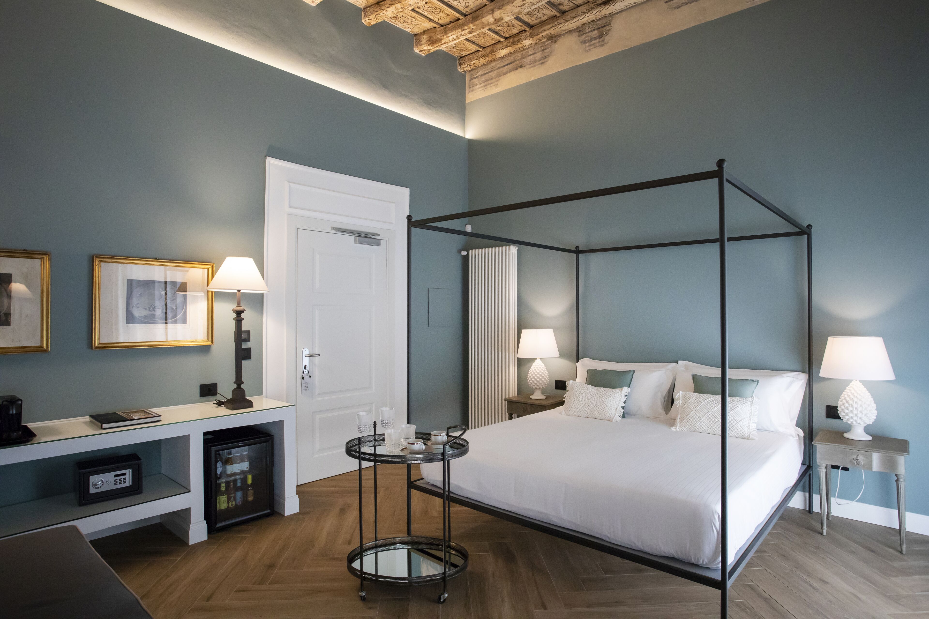 Una habitación de hotel moderna con una cama con dosel, un escritorio con ordenador, un refrigerador pequeño y una mesita de noche con lámpara.