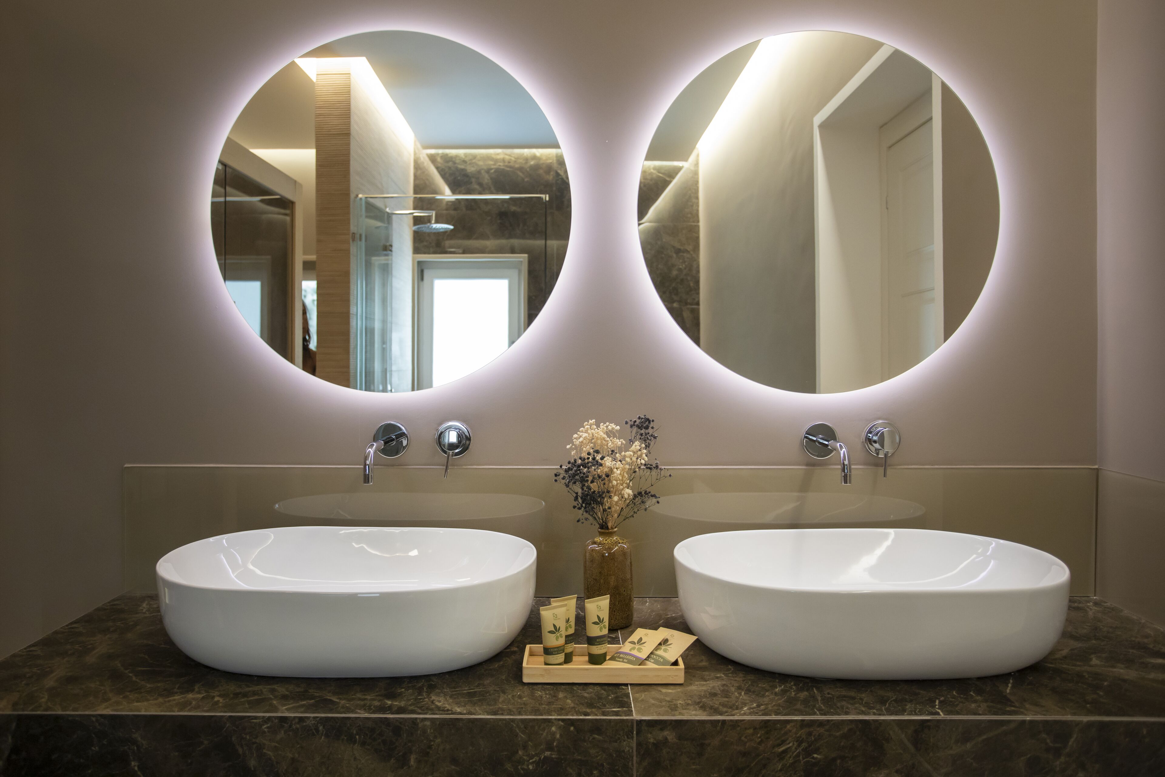 Une salle de bain avec deux grands lavabos ovales, un miroir au-dessus, et un petit vase avec des fleurs posé sur le comptoir.