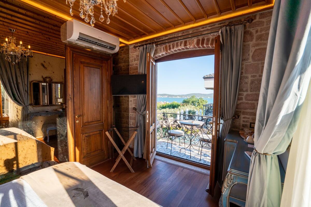 A room with a wooden ceiling, an air conditioning unit, a chandelier, a wooden door, a small table with chairs, and a view of a balcony with outdoor furniture.