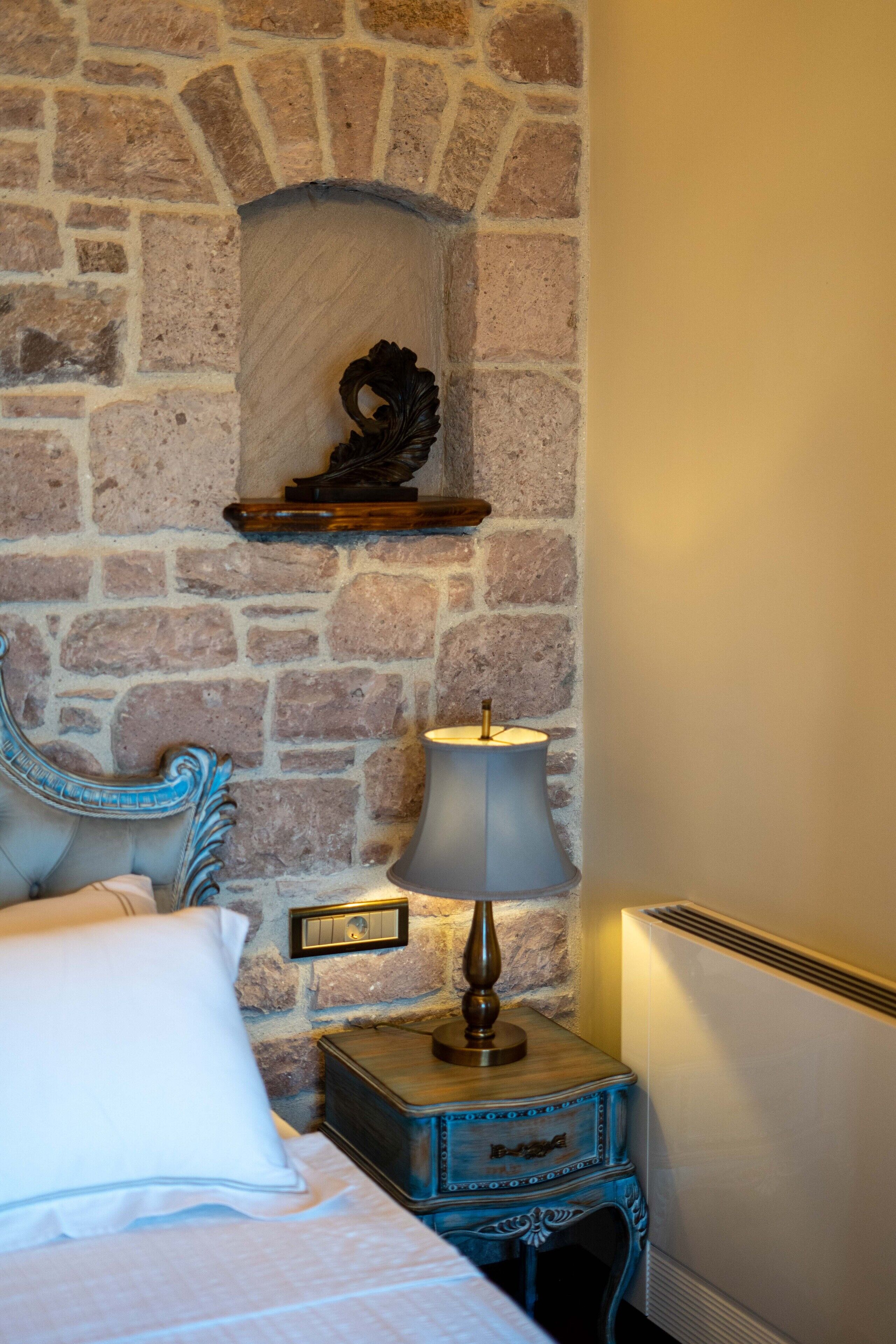 Una habitación con pared de piedra, una cama con cabecero azul, una mesita de noche con una lámpara y un pequeño estante sobre la pared.