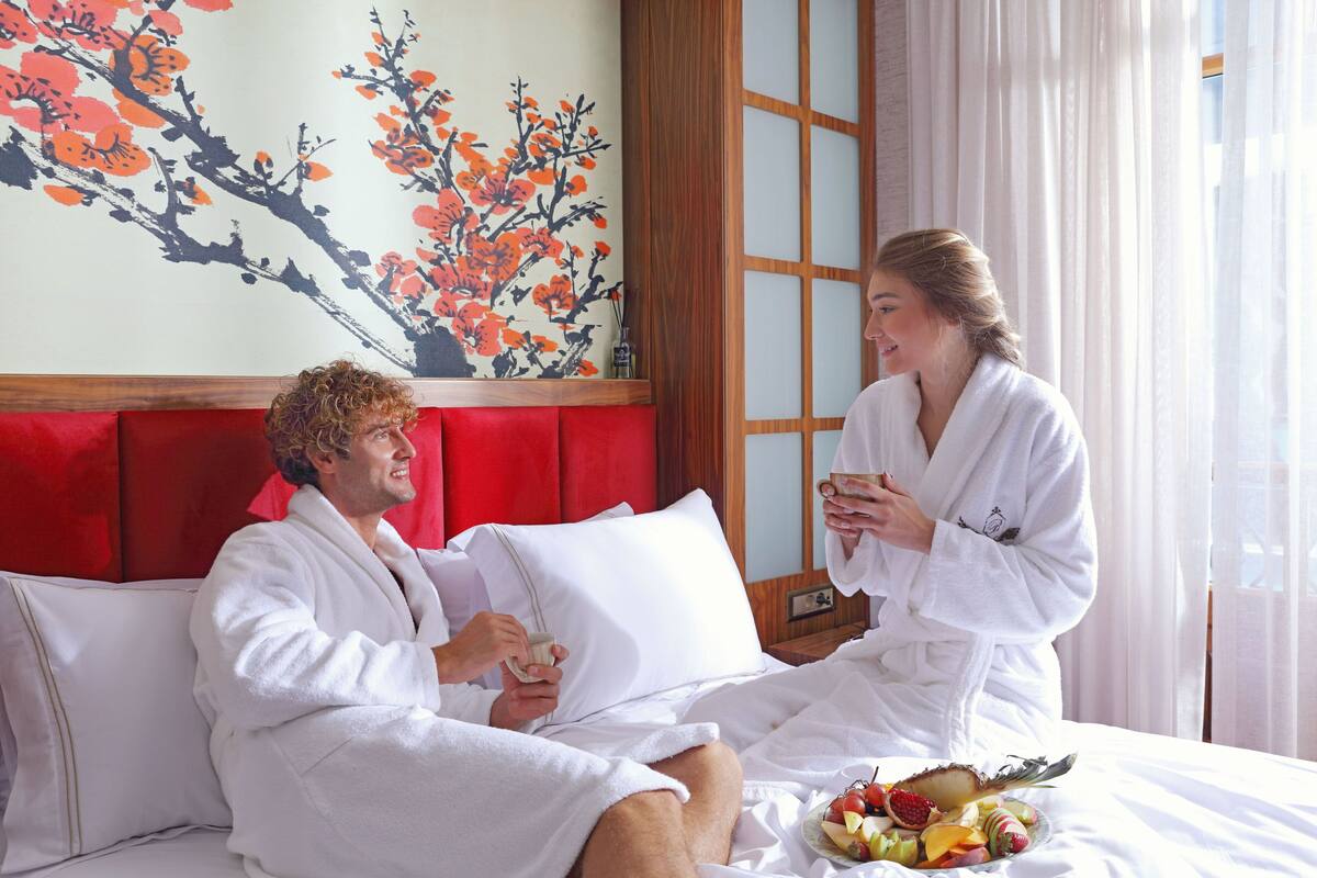 A hotel room with a red headboard, a painting of a tree with orange flowers, and a fruit basket on the bed.