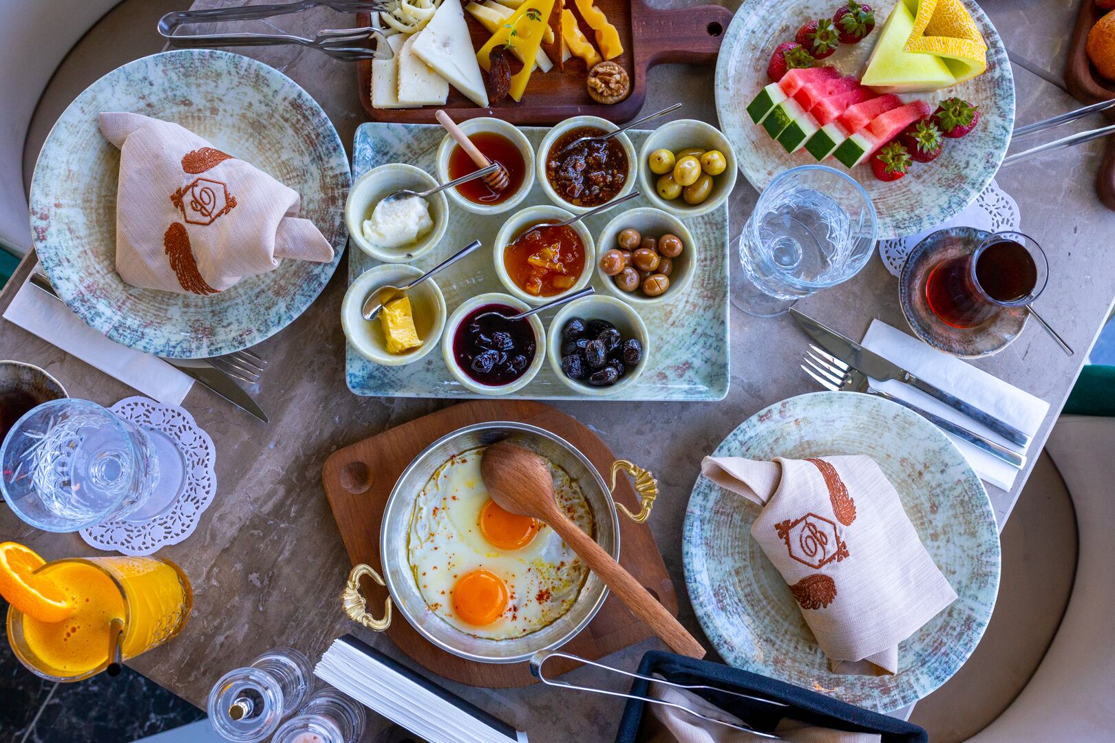 A breakfast setting with plates of eggs, cheese, and fruits, along with various condiments and a glass of orange juice.