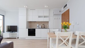 A modern kitchen with white cabinets, a built-in oven, and a coffee maker. There is a dining area with white chairs and a wooden table. - Intimate Apartment | SPA & Gym | 550 m to the beach (Miedzywodzie)