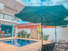 Piscina con deck de baldosas, un paraguas verde y mobiliario exterior.
