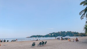 A beach with rows of green beach chairs and umbrellas, a sandy shore, and a hill with buildings in the background. - Pousada BellaSophia (Guarujá)