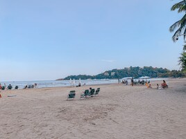 Una playa con hileras de sillas y sombrillas verdes, una orilla arenosa y una colina con edificios al fondo.