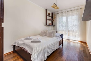 A bedroom with a bed, wooden flooring, a ceiling light fixture, a window with sheer curtains, and a wall-mounted shelf with decorative items.