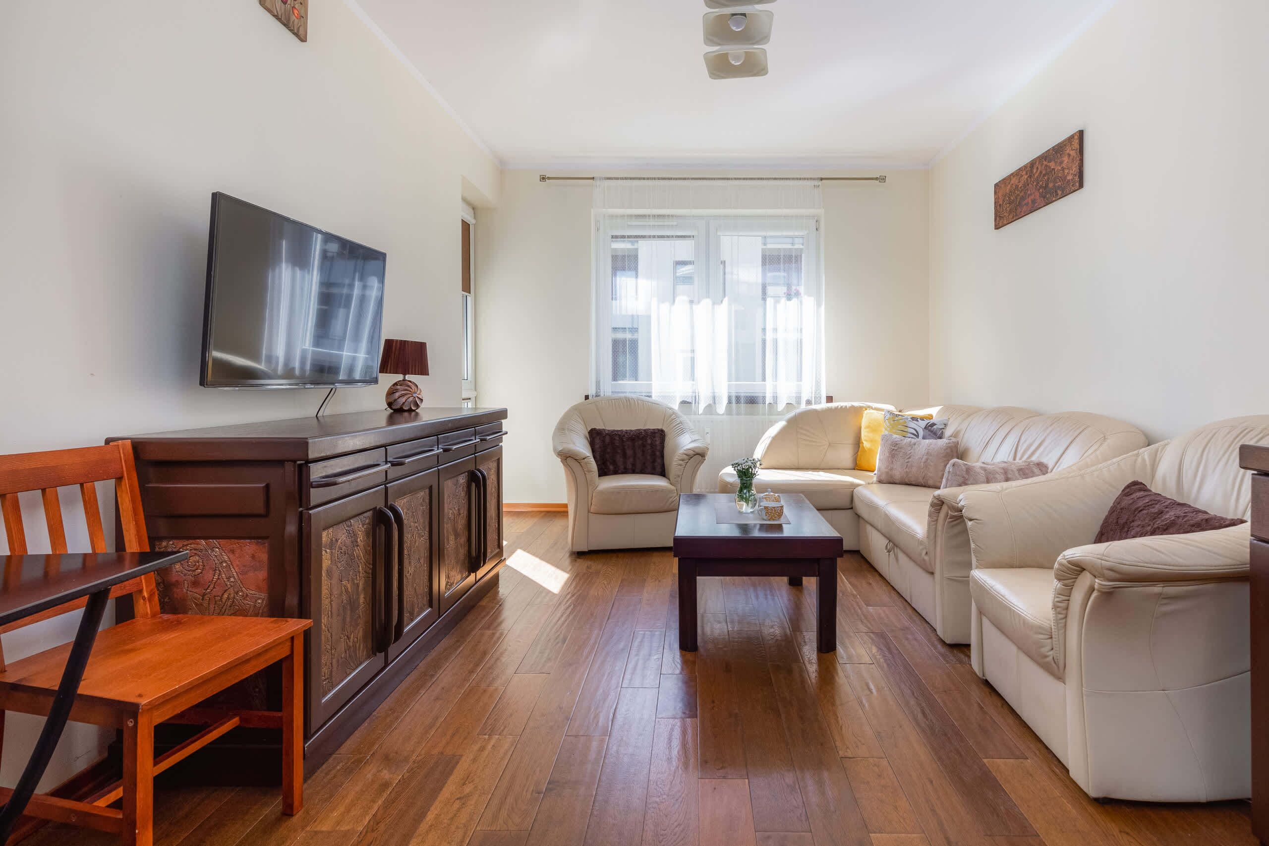 Una sala de estar con televisor de pantalla plana, muebles de madera, un conjunto de sofás y un ventanal con cortinas.