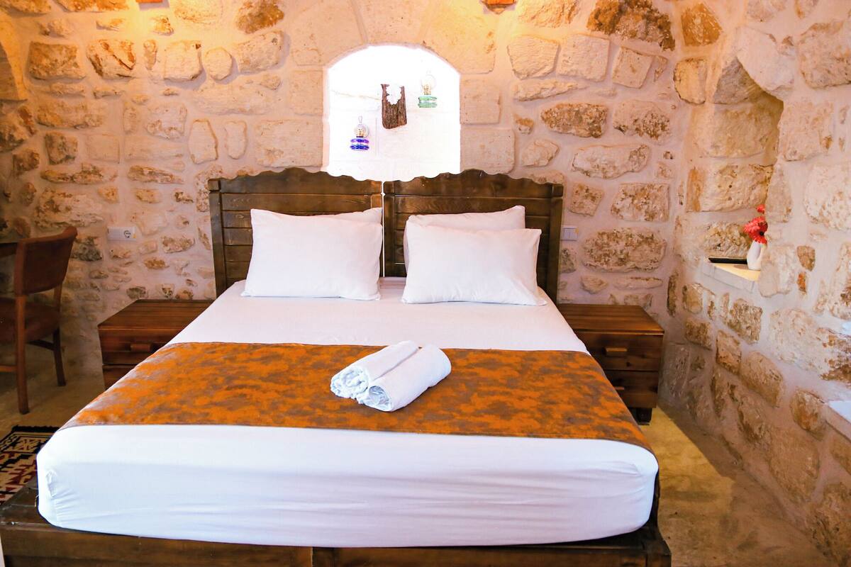 A room with a stone wall, a wooden bed with white bedding, and a small window with a view of the outside.