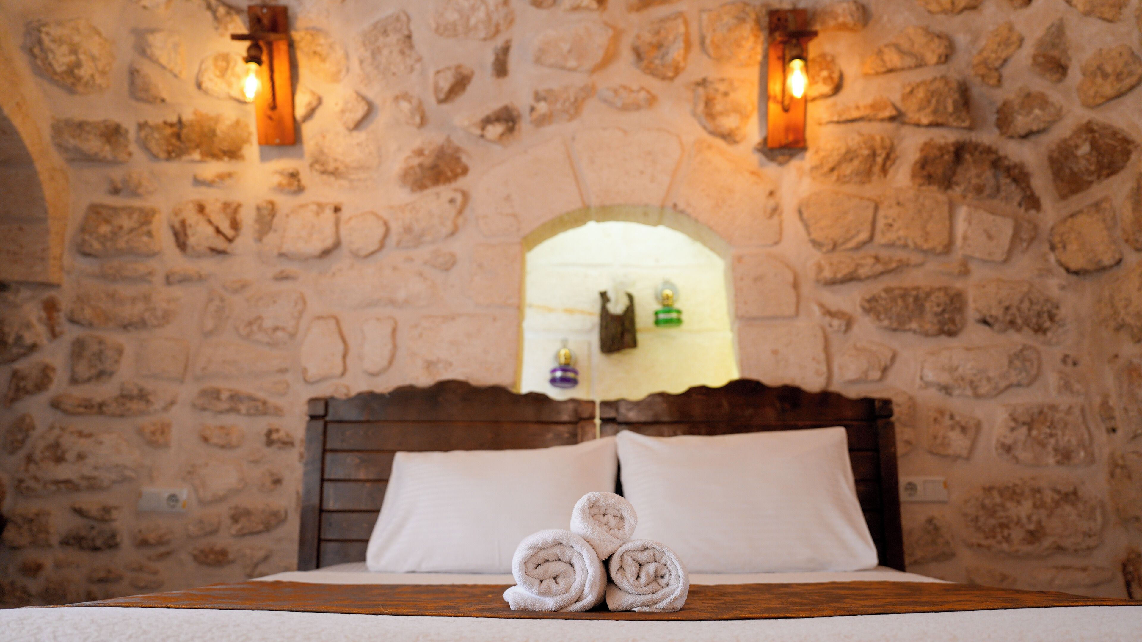 A stone-walled room with a wooden bed, two wall-mounted lights, and a mirror reflecting a green and purple object.