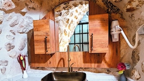 Un baño con pared de piedra, armarios de madera, un lavabo con pileta decorativa y una ventana con cortina.