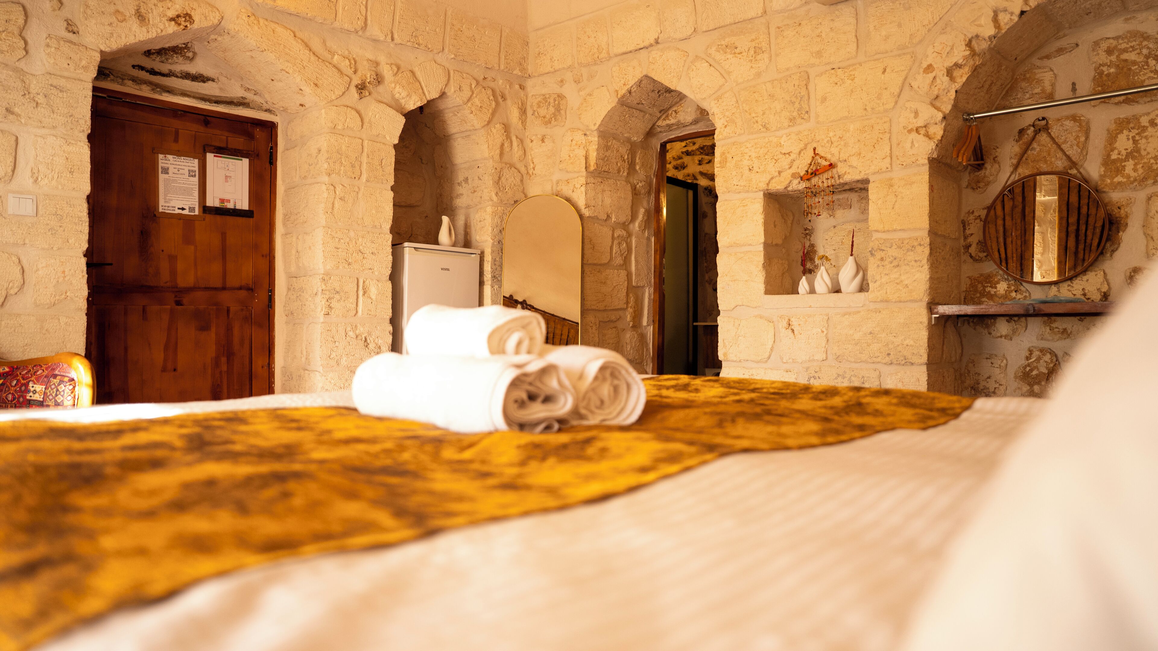 A room with stone walls, a wooden door, a bed with a yellow blanket, and a mirror on the wall.