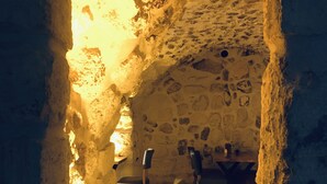 Un interior rústico de piedra con una mesa de madera, dos faroles y un jarrón con plantas secas.