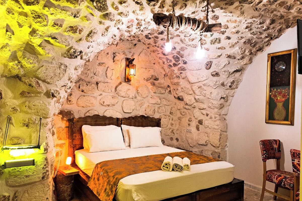 A stone-walled bedroom with a wooden bed, a nightstand, a chair, and a painting on the wall.