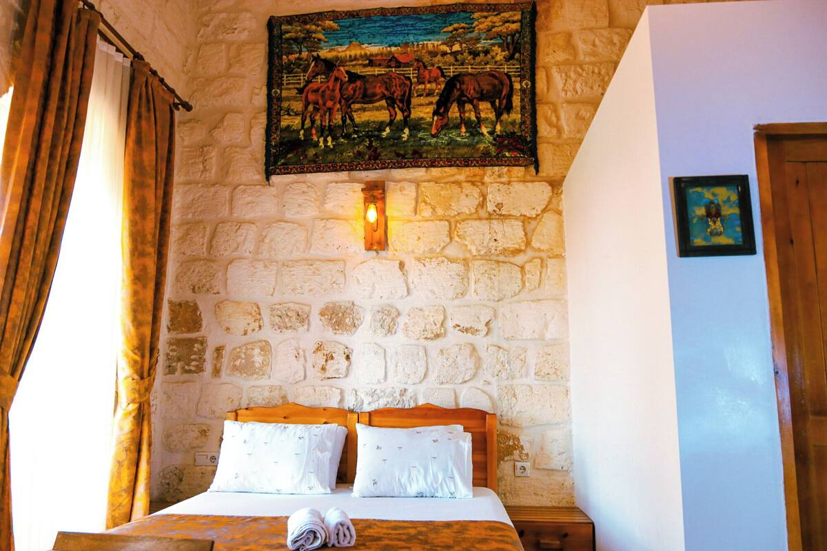 A room with a stone wall, a bed with two pillows, a wooden headboard, a wooden nightstand, a wall-mounted light, and a painting of horses.