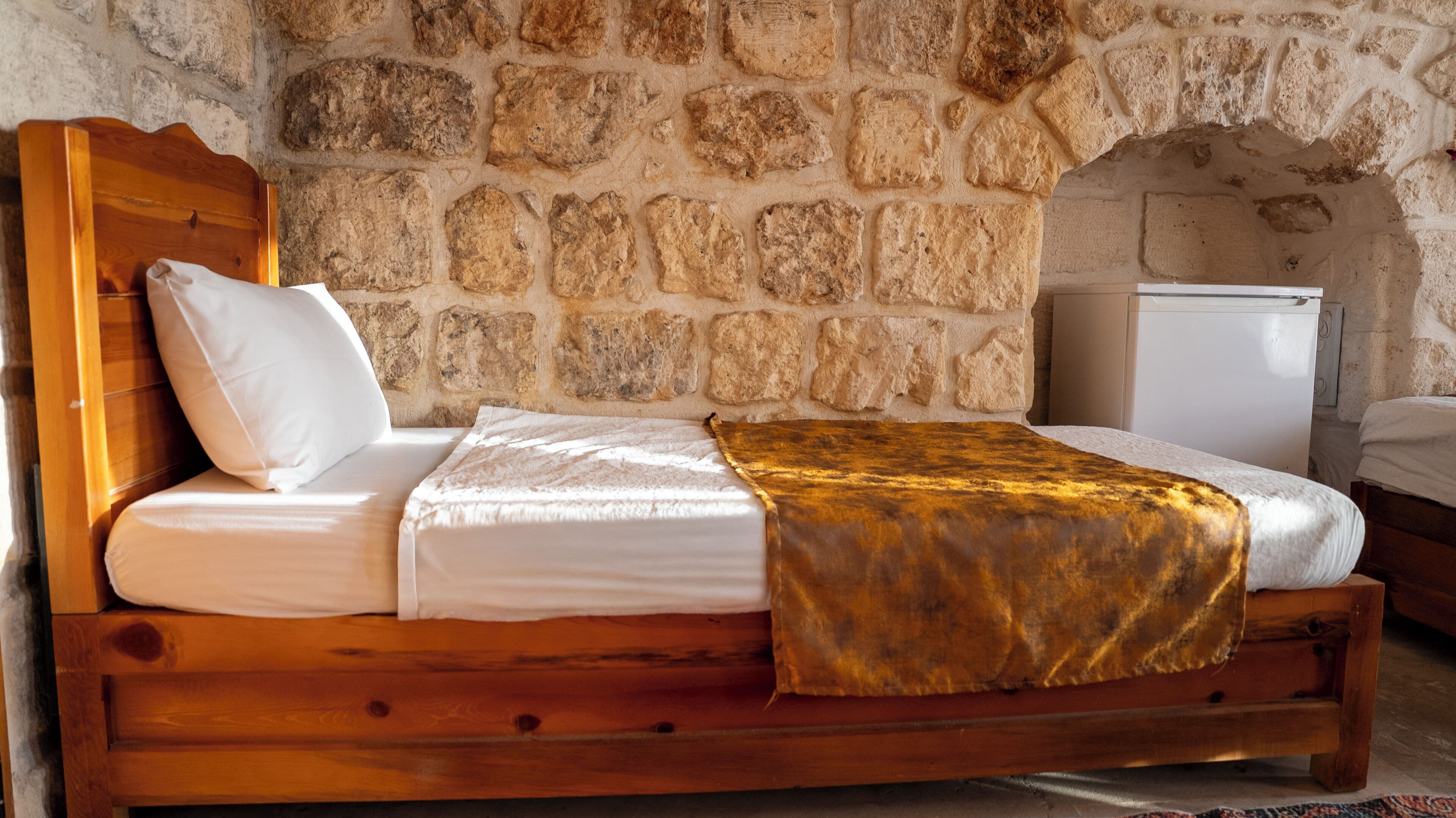 Un letto in legno con lenzuola bianche e una coperta in oro, addossato a un muro di pietra con una nicchia ad arco.