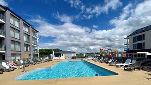 Ein Schwimmbecken mit Liegestühlen, ein Spielbereich für Kinder und moderne Gebäude mit Balkonen.