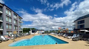 Una piscina con sillas de descanso, un área de juegos para niños y un edificio con balcones.