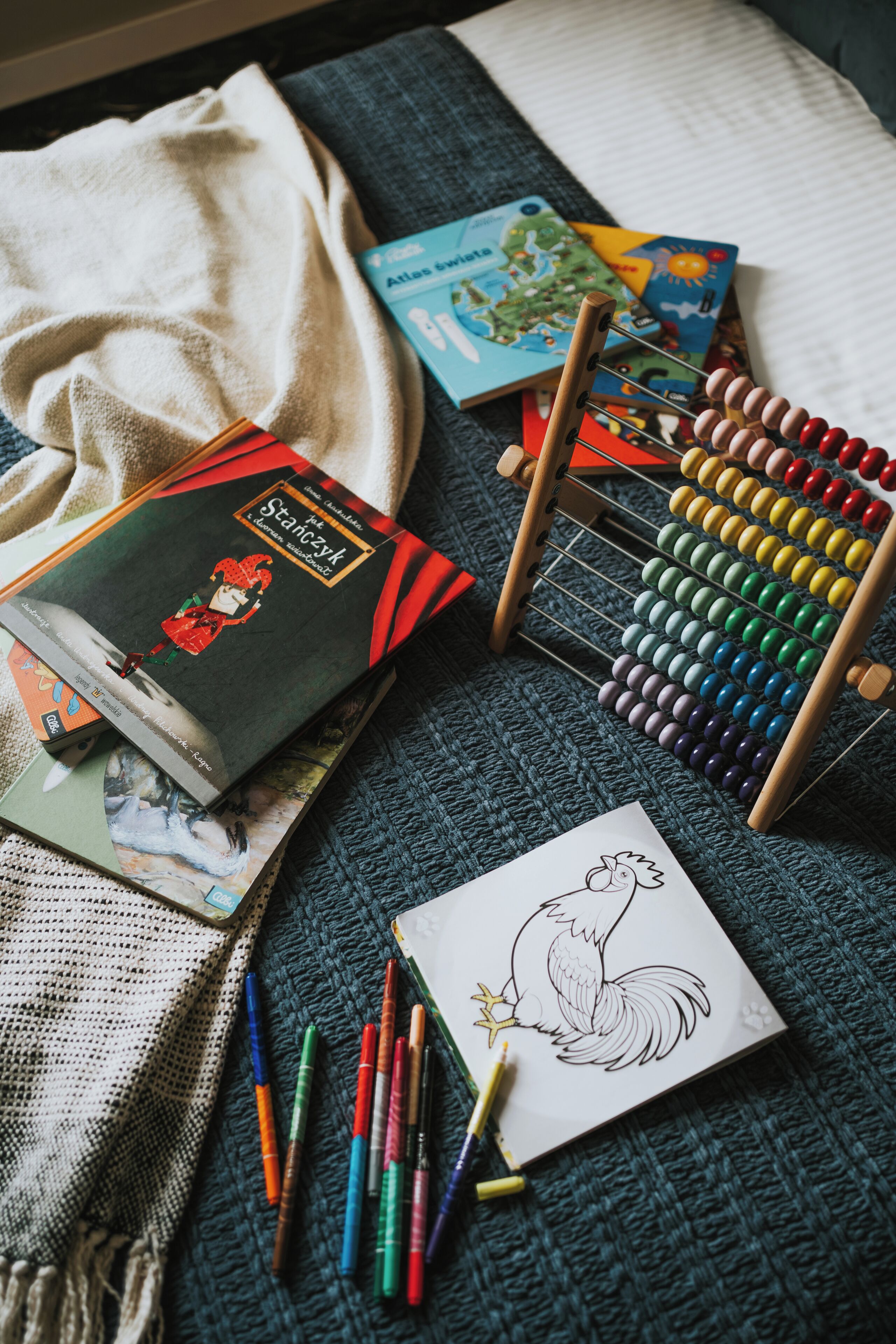 A book titled "Staszysz" on a blue knitted blanket, a wooden abacus, and a drawing of a rooster with colored pencils.