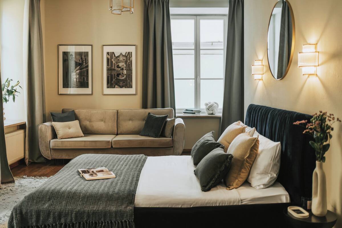 Een slaapkamer met een bed, een bank, een raam met gordijnen, ingelijste foto’s aan de muur en wandlampen.