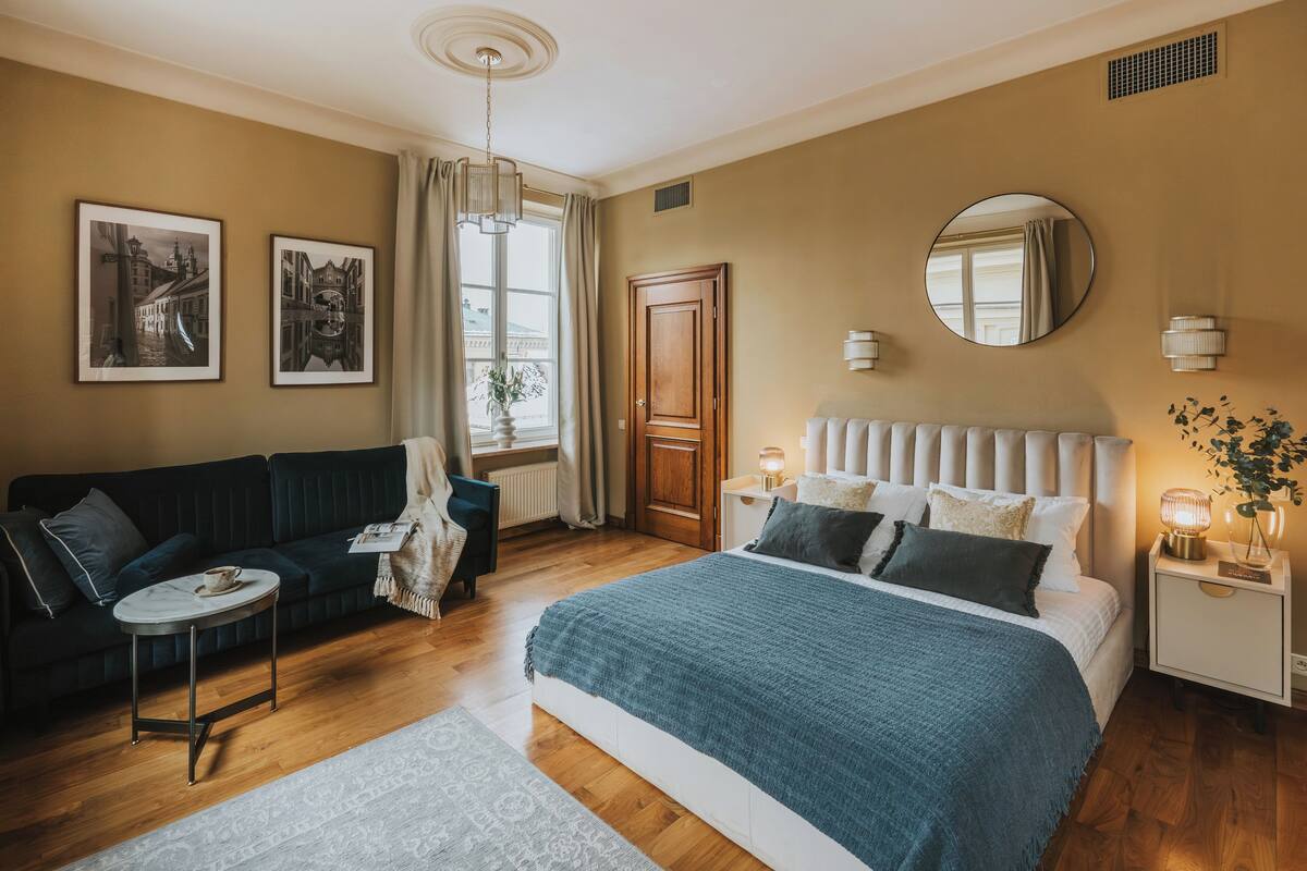 Een slaapkamer met een bed, een bank, een klein tafeltje, een spiegel en een raam met gordijnen.