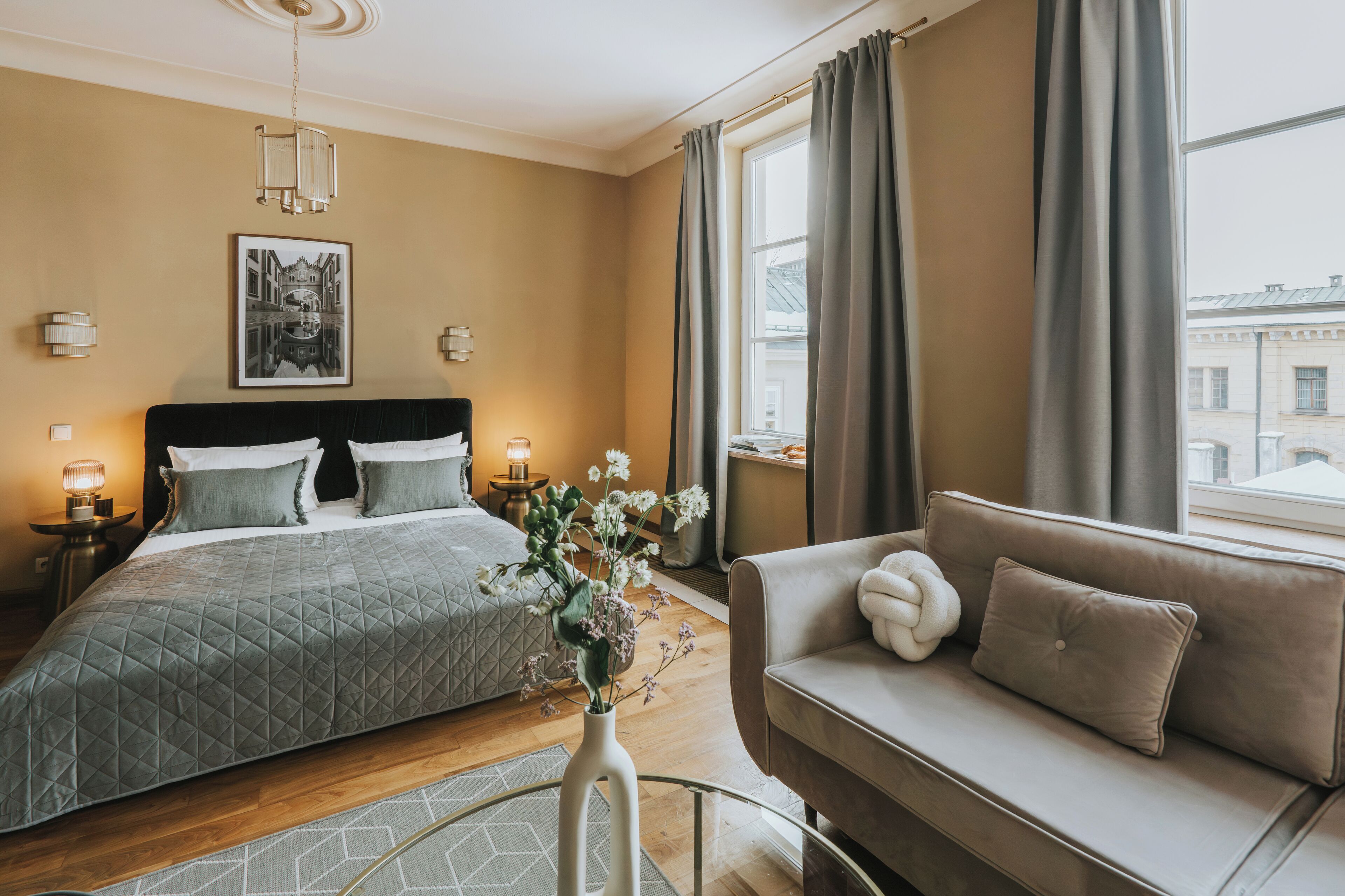 Une chambre à coucher avec un lit, un canapé, un vase de fleurs, un tableau encadré au mur et une fenêtre avec des rideaux.