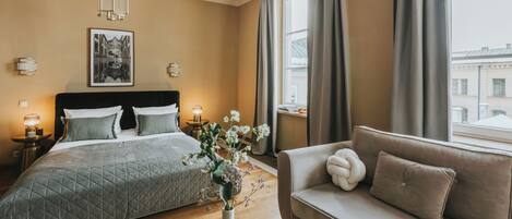 A bedroom with a bed, a sofa, a vase with flowers, a framed picture on the wall, and a window with curtains.