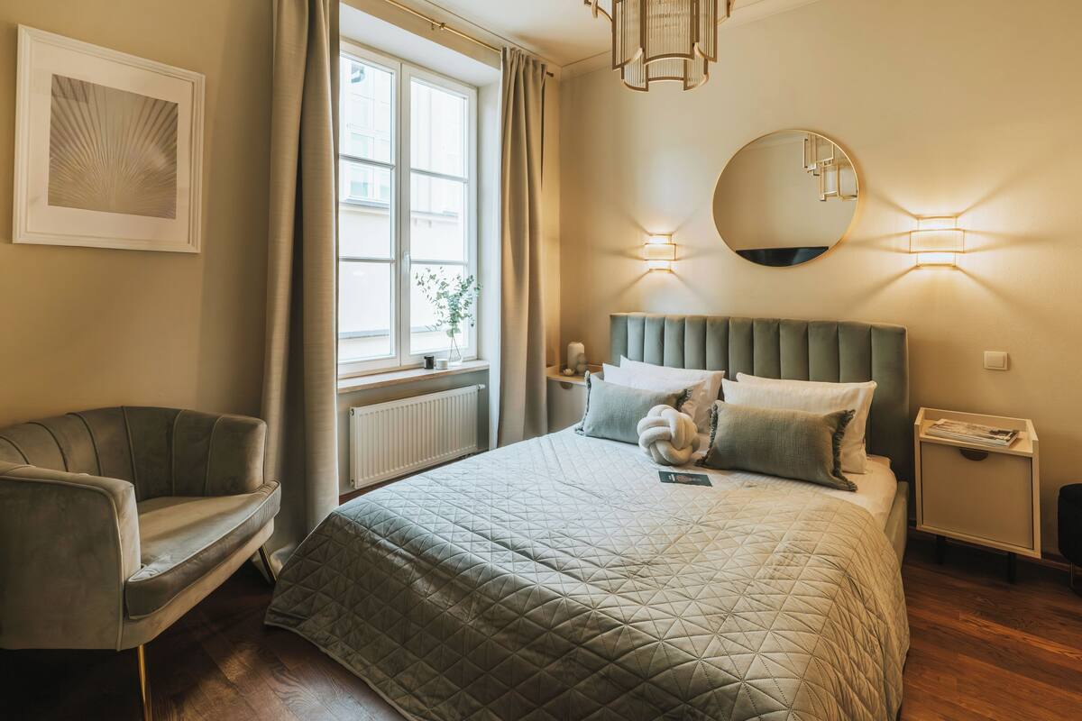 Een keurig opgemaakt slaapkamer met een groot bed, een stoel, een raam met gordijnen, een ronde spiegel en een nachtkastje.