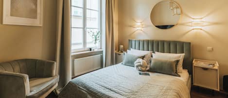 A neatly arranged bedroom with a large bed, a chair, a window with curtains, a round mirror, and a bedside table.