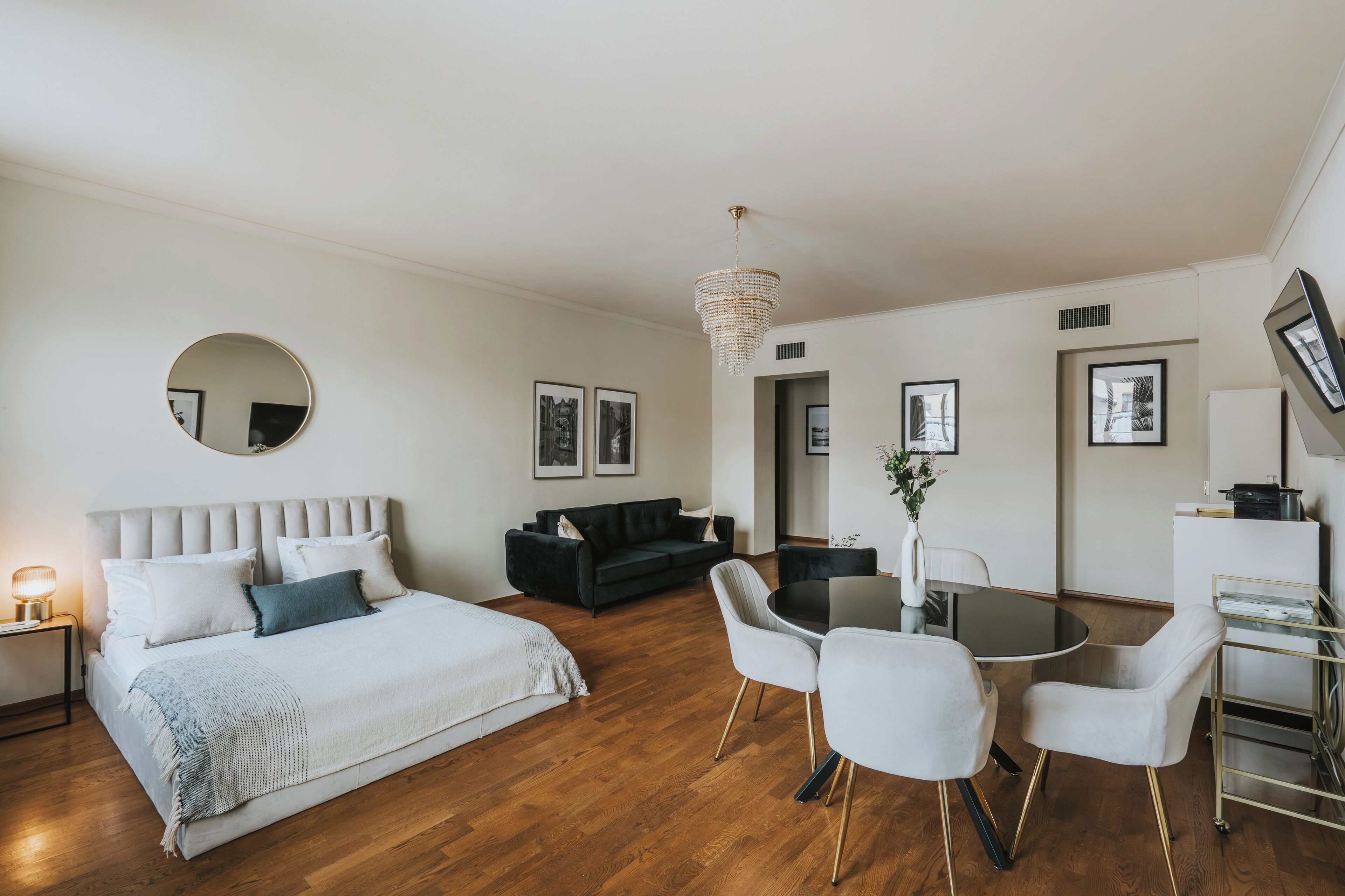 A modern hotel room with a bed, a dining table with chairs, a small kitchen area, and a living space with a sofa.