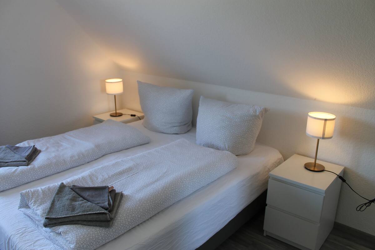 A neatly made twin bed with a white duvet and two pillows, a bedside table with a lamp, and a small drawer unit.