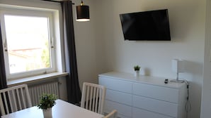 Una habitación con una mesa blanca, sillas, una planta en maceta, un armario blanco, un televisor montado en la pared y una ventana con cortinas.