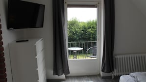 Un dormitorio con televisor, cama, radiador, ventana con cortinas y balcón con mesa y silla.