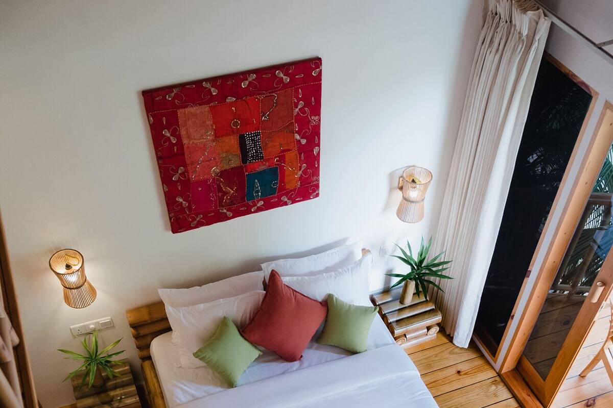 Una cama con estructura de madera, ropa de cama blanca y una manta roja. Encima de la cama, hay un colcha roja y azul en la pared.
