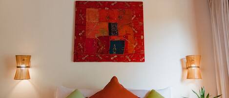 A bed with a red bedspread, multiple pillows, and a red abstract painting on the wall. There are two wall-mounted lamps on either side of the bed.