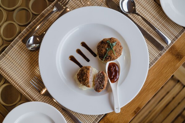 A white plate with two round, breaded items and a spoon with sauce on a checkered tablecloth. - Ecoboo Maldives (Thinadhoo)