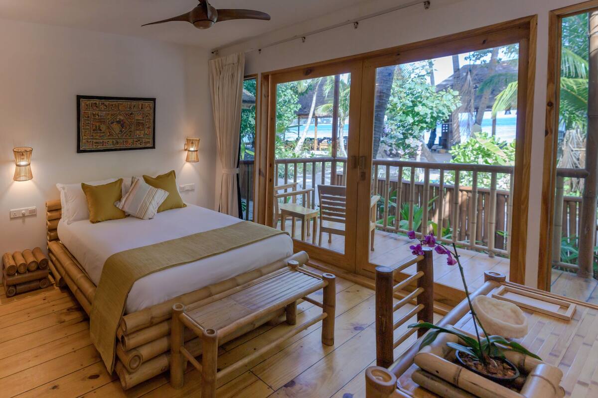 Una habitación con cama, piso de madera, ventilador de techo, ventana grande y balcón con vistas al exterior.