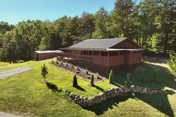 A wooden cabin with a sloped roof, surrounded by a stone wall and a well-maintained lawn with small trees.