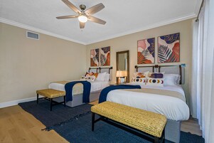 A hotel room with two beds, a ceiling fan, artwork on the walls, and a woven bench.