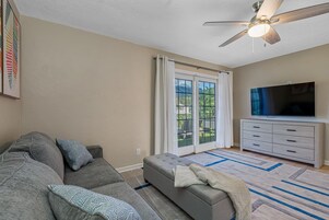 A living room with a grey sofa, a white ottoman, a flat-screen TV, and a ceiling fan.