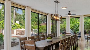 A spacious dining area with a long wooden table, multiple chairs, and a central decorative bowl. Large windows provide a view of a lush garden and outdoor seating area. - 8 BR Heated Pool & Soccer Field (Largo)