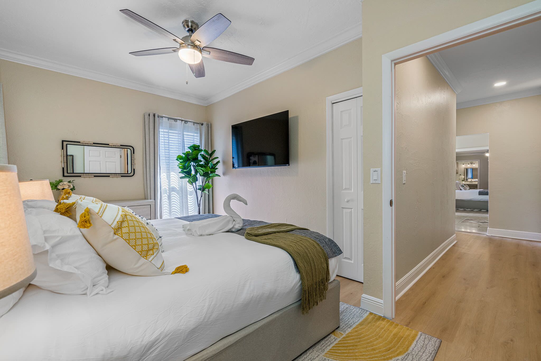 A bedroom with a bed, a ceiling fan, a TV mounted on the wall, and a door leading to another room.