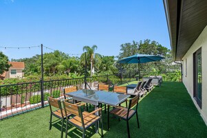 Un patio al aire libre con una mesa de vidrio y sillas de madera, rodeado por una valla de metal y abundante vegetación.