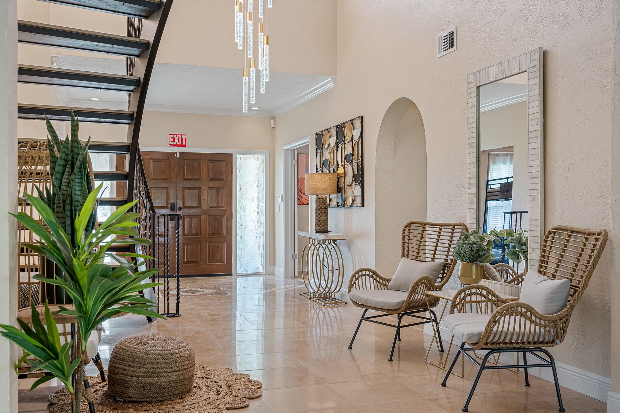 A spacious lobby with a staircase, wicker chairs, a large mirror, and a decorative wall piece.