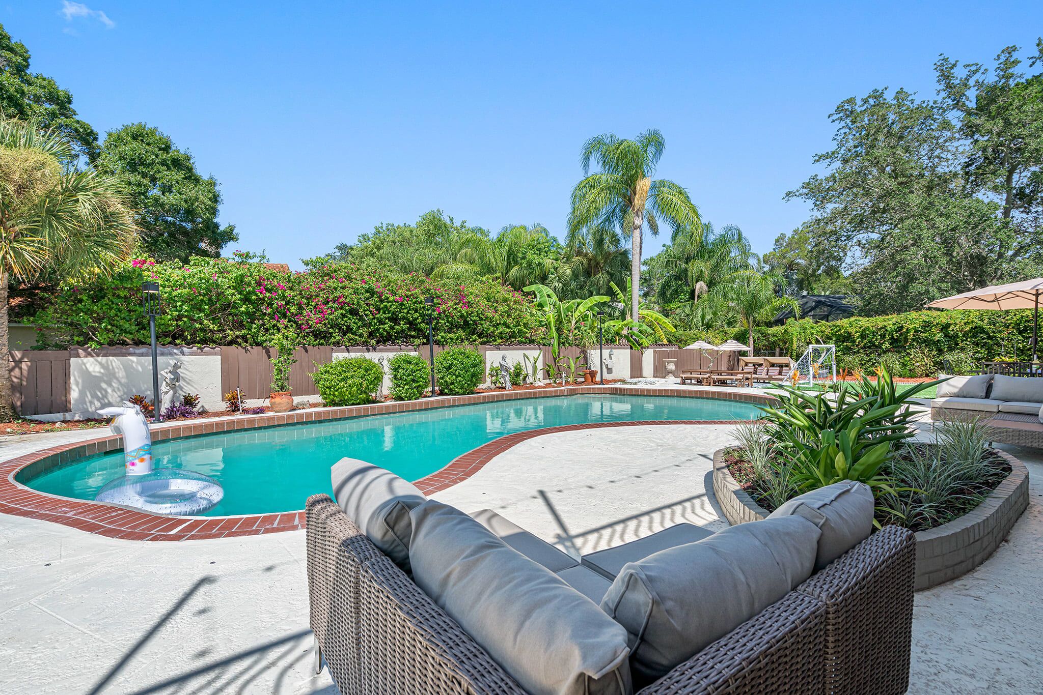 A swimming pool with a brick edge, surrounded by a garden with various plants and a wicker lounge area.