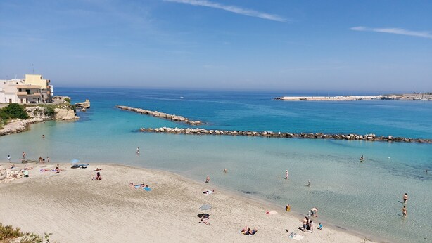 Ein Strand mit klarem Wasser, einem Felssteg und Gebäuden im Hintergrund.