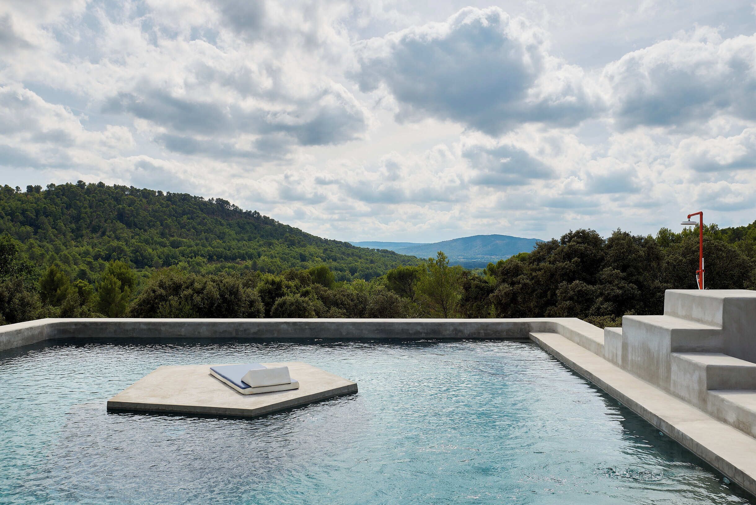 A swimming pool with a floating platform and steps leading into the water, surrounded by lush greenery and hills.