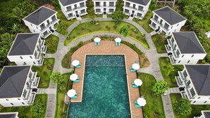 Une vue aérienne d’un complexe hôtelier avec une piscine centrale entourée de chaises longues et de parasols, ainsi que plusieurs villas à deux étages disposées autour de la piscine.