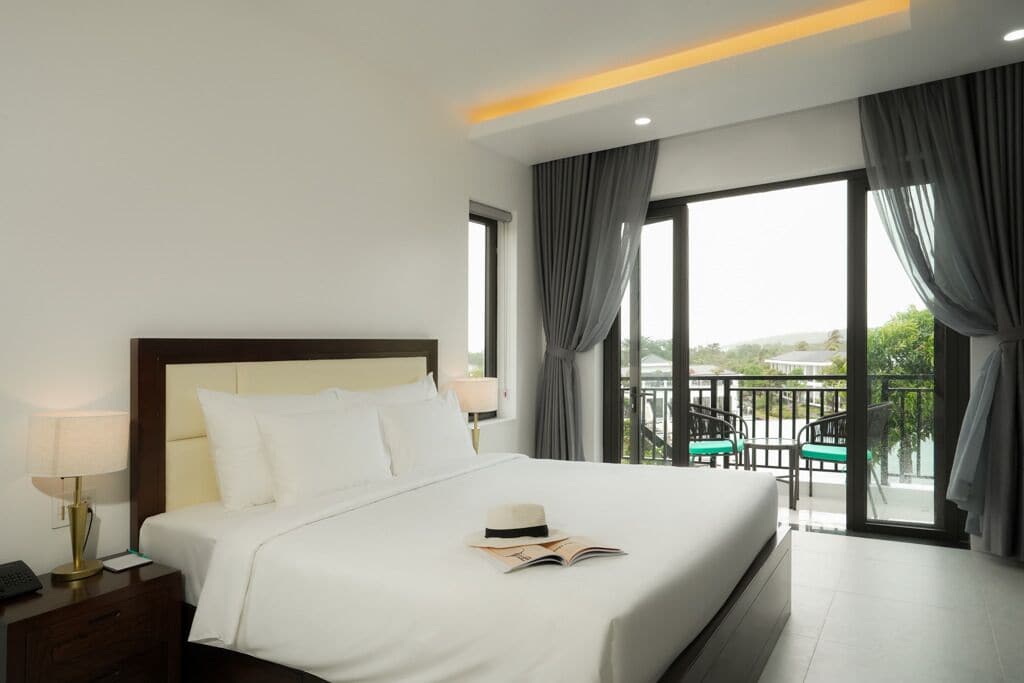 Habitación de hotel con una cama grande, una mesita de noche con lámpara y vistas al exterior a través de amplias ventanas.