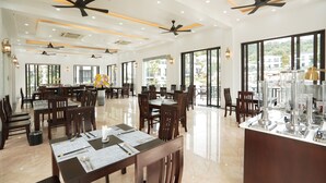 Un vaste espace dédié à la salle à manger, avec de nombreuses tables et chaises, des ventilateurs de plafond et de grandes fenêtres offrant une vue sur l’extérieur.